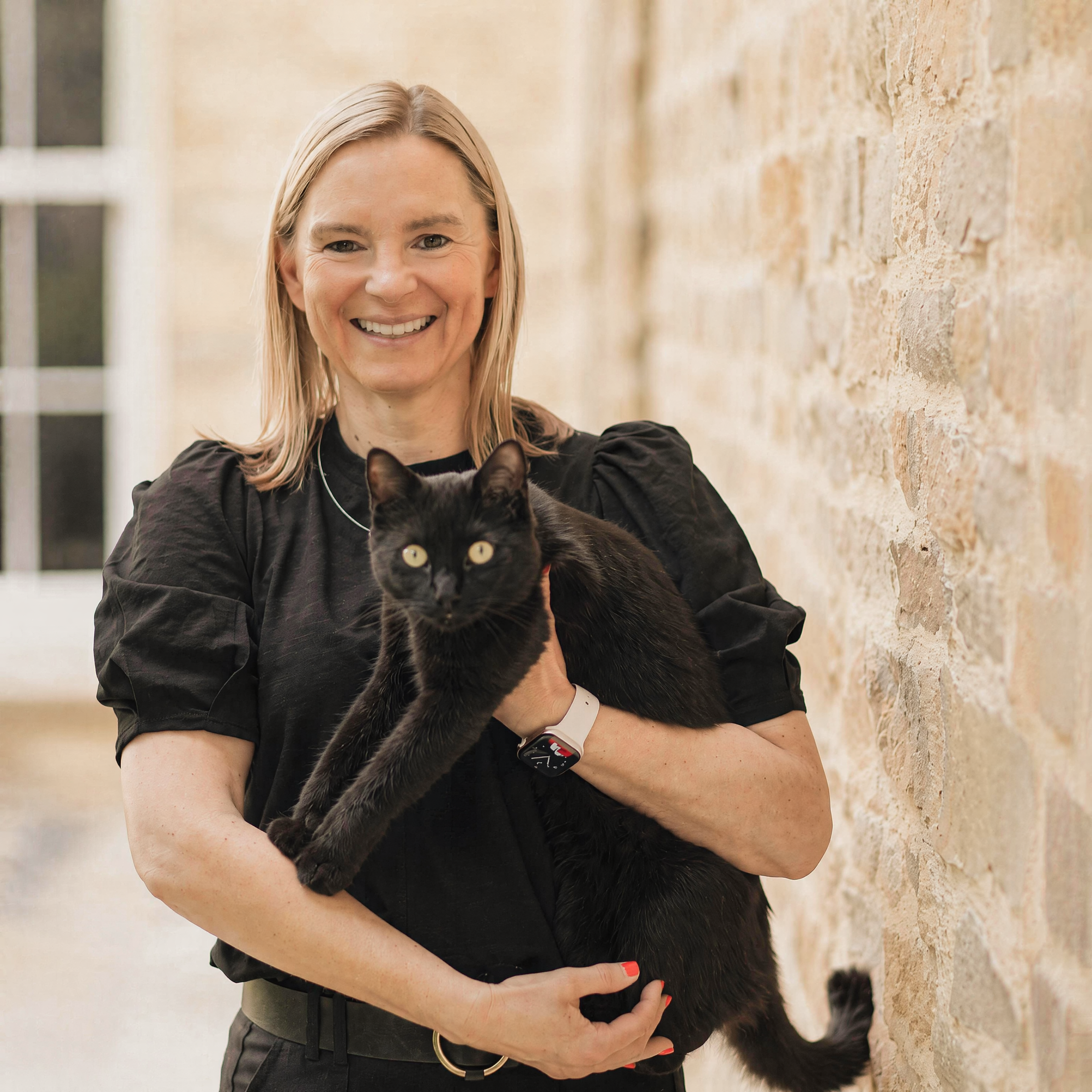 Sam Bowman smiling while cradling her black cat, leaning against a warm stone wall outdoors.