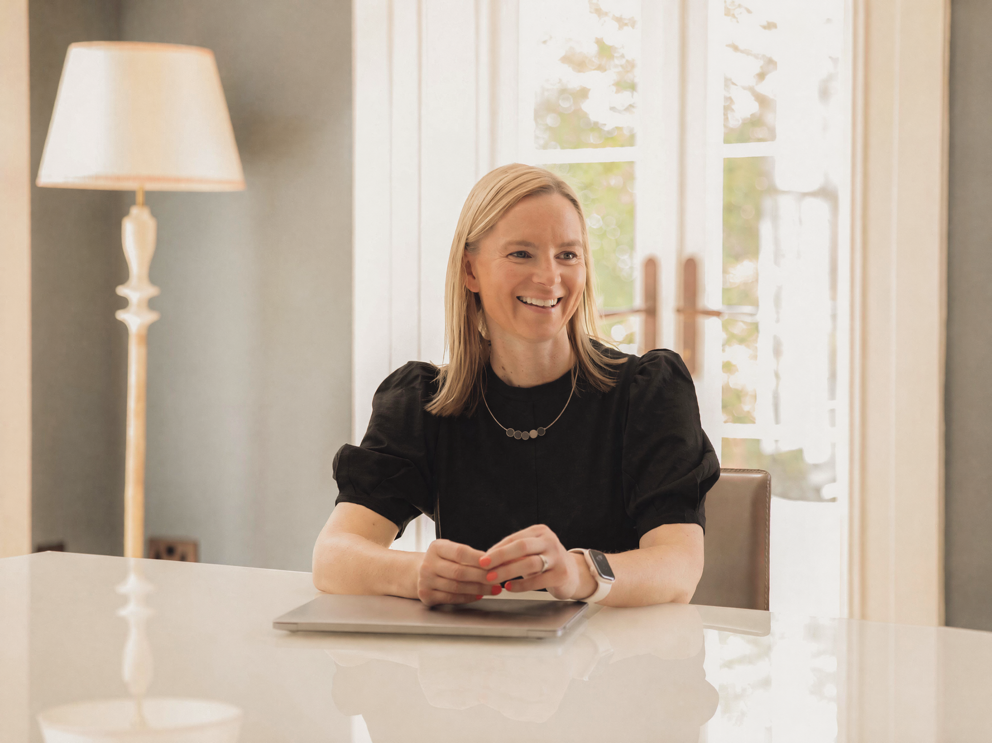 Sam Bowman, Talkabout's Clinical Director, smiling at a sunlit table with a closed laptop in a calm, light-filled room.